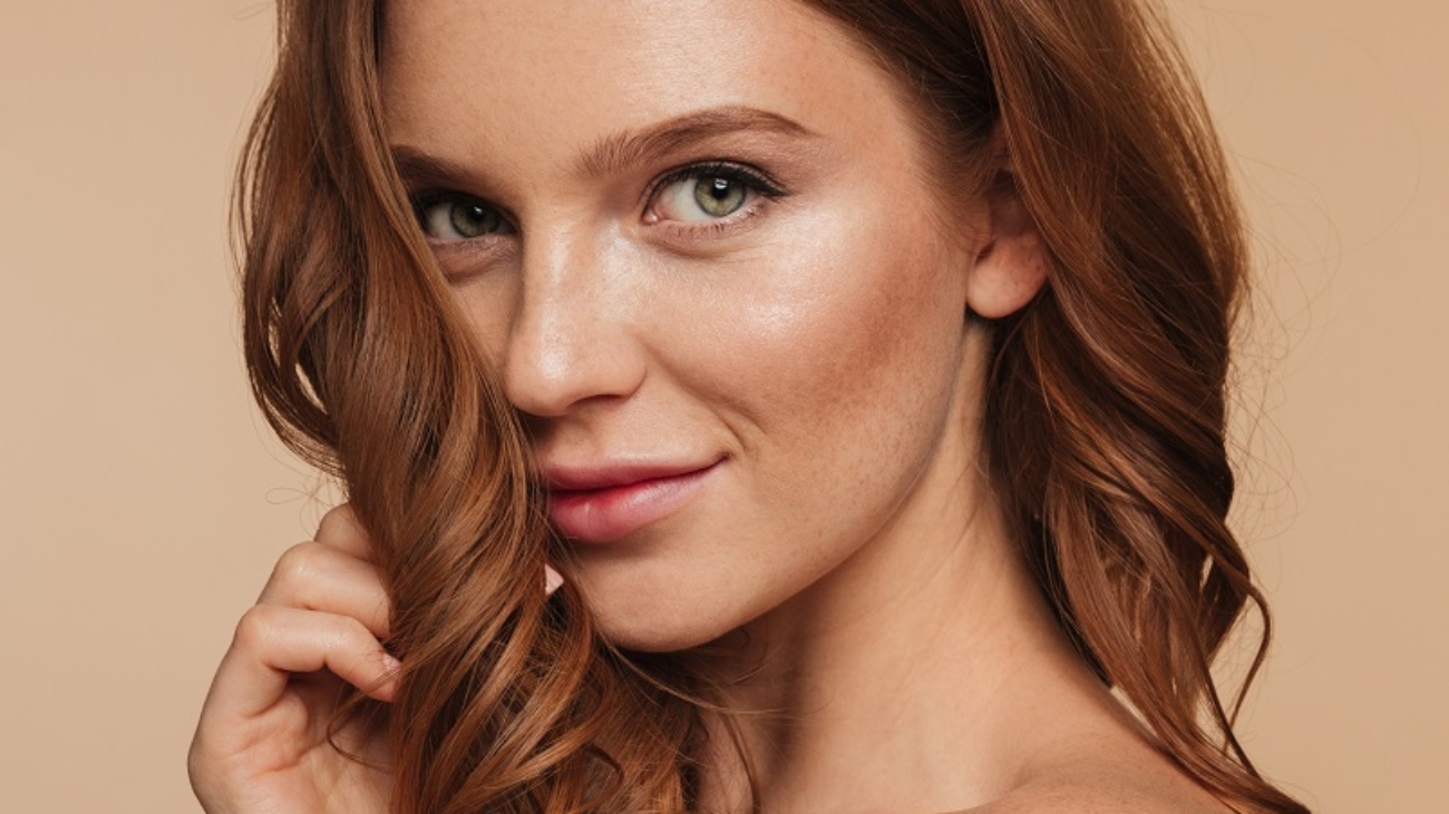 Beauty portrait of mystery smiling ginger woman with long hair posing sideways and looking at the camera over cream background