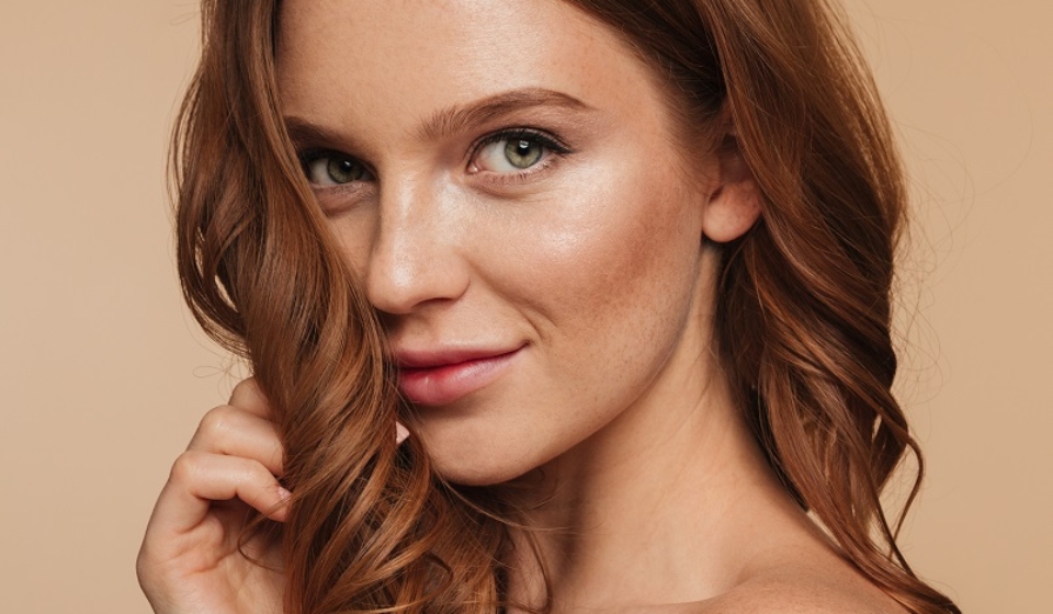 Beauty portrait of mystery smiling ginger woman with long hair posing sideways and looking at the camera over cream background