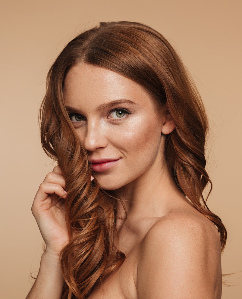 Beauty portrait of mystery smiling ginger woman with long hair posing sideways and looking at the camera over cream background