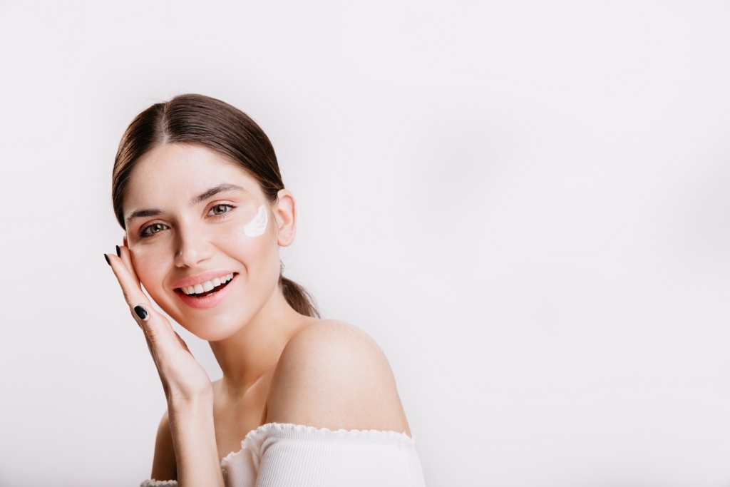 Girl touches moisturized skin and smiles. Portrait of model with cream on face on isolated background