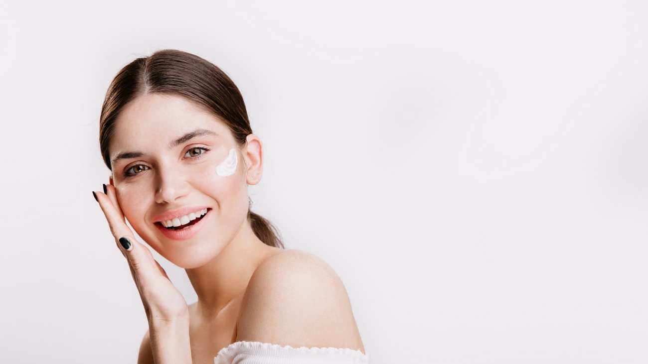 Girl touches moisturized skin and smiles. Portrait of model with cream on face on isolated background