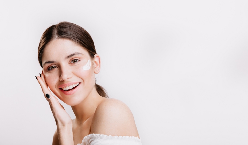 Girl touches moisturized skin and smiles. Portrait of model with cream on face on isolated background